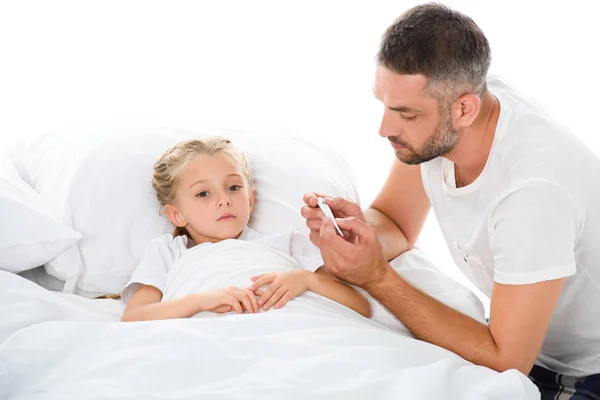 Padre mirando termómetro electrónico mientras hija enferma con temperatura acostada en la cama, aislada en blanco - foto de stock
