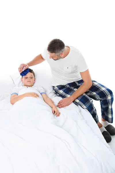 Батько тримає крижаний пакет на голові хворого сина з температурою, що лежить у ліжку, ізольований на білому — стокове фото