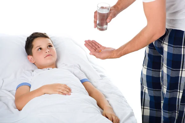 Батько дає таблетки і склянку води хворим сином в ліжку, ізольовані на білому — стокове фото