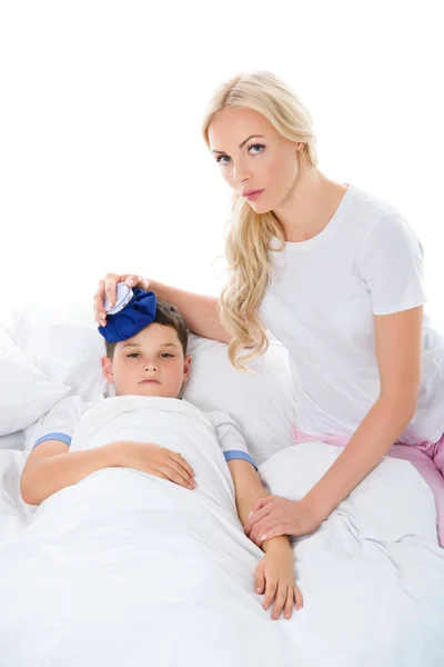 Madre sosteniendo bolsa de hielo en la cabeza del hijo enfermo con temperatura acostado en la cama, aislado en blanco - foto de stock