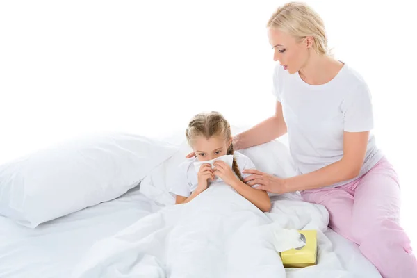Malade fille soufflant son nez liquide dans le lit tandis que maman assis près, isolé sur blanc — Photo de stock