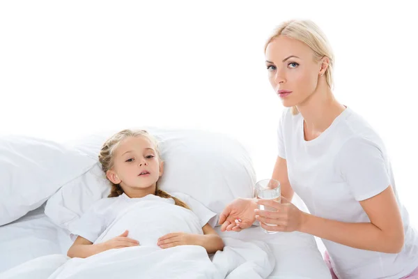 Мать дает таблетки и стакан воды больной дочери в постели, изолированные на белом — стоковое фото