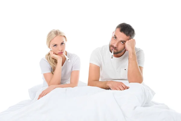 Casal doente com termômetros eletrônicos sentados na cama, isolados em branco — Fotografia de Stock