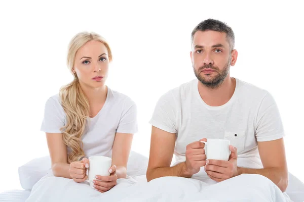 Pareja enferma sentada en la cama con tazas de bebida caliente, aislada en blanco - foto de stock