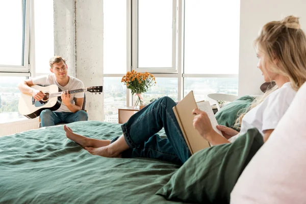 Mujer joven con libro descansando en la cama mientras su novio toca la guitarra acústica en casa - foto de stock