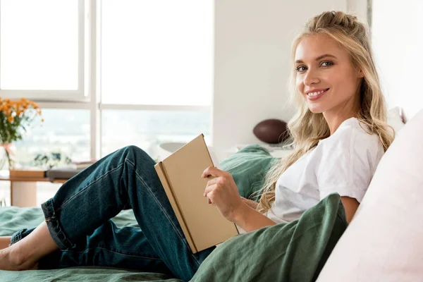 Vista lateral da jovem mulher bonita com livro descansando na cama em casa — Fotografia de Stock