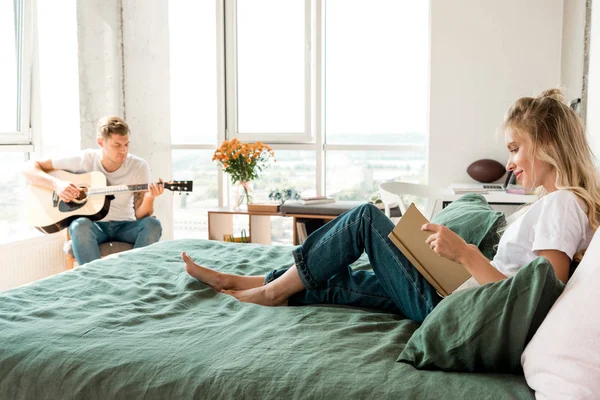 Jeune femme lecture livre sur lit tandis que copain jouer de la guitare acoustique à la maison — Photo de stock
