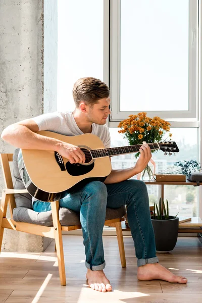 Joven en sillón tocando la guitarra acústica en casa - foto de stock