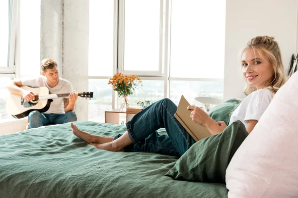 Mujer joven con libro descansando en la cama mientras su novio toca la guitarra acústica en casa - foto de stock