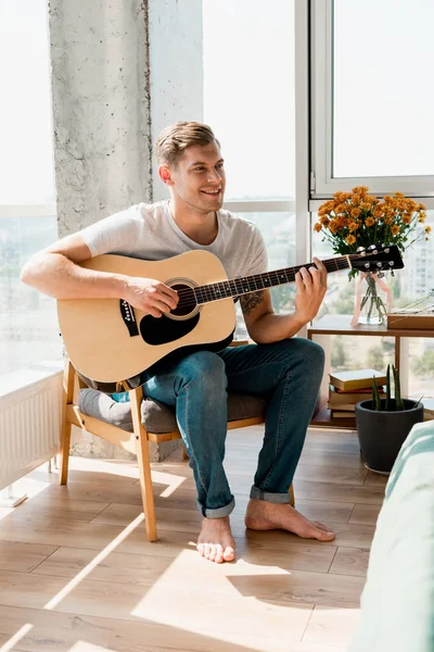 Giovane uomo sorridente in poltrona che suona la chitarra acustica a casa — Foto stock