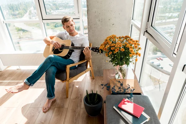 Junger Gitarrist spielt Akustikgitarre, während er zu Hause im Sessel liegt — Stockfoto