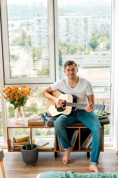 Joven guitarrista tocando la guitarra acústica mientras está sentado en la ventana en casa - foto de stock
