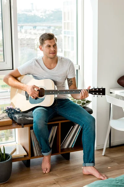 Joven guitarrista masculino pensativo en ropa casual tocando la guitarra acústica en casa - foto de stock