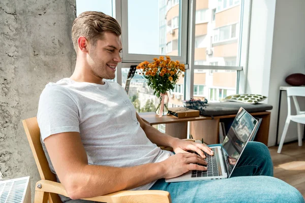 Uomo sorridente in poltrona utilizzando il computer portatile a casa — Foto stock