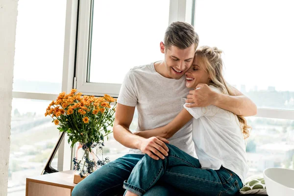 Heureux jeune couple en amour dans des vêtements décontractés étreignant à la fenêtre à la maison — Photo de stock