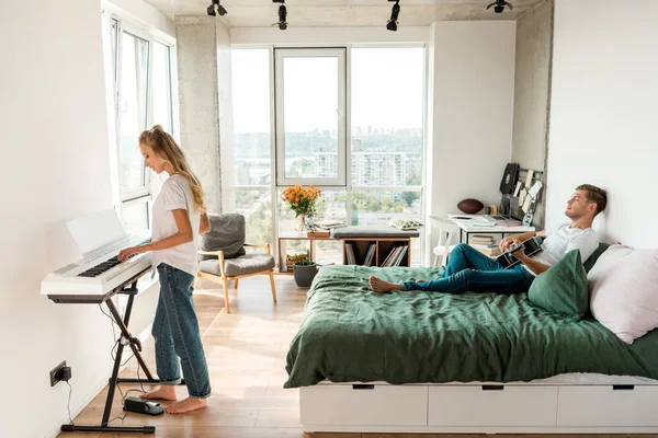 Vista lateral del hombre tocando la guitarra acústica en la cama mientras su novia toca el piano electrónico en casa - foto de stock