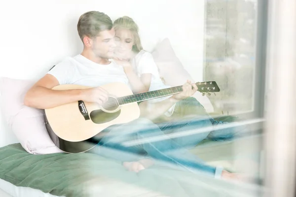 Мужчина играет на акустической гитаре для подружки, отдыхая дома на кровати — стоковое фото