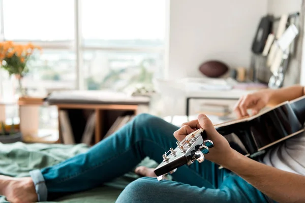 Tiro recortado do guitarrista em jeans tocando guitarra acústica na cama em casa — Fotografia de Stock