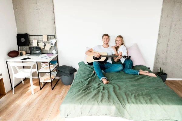 Hombre tocando la guitarra acústica para novia mientras descansa en la cama en casa - foto de stock