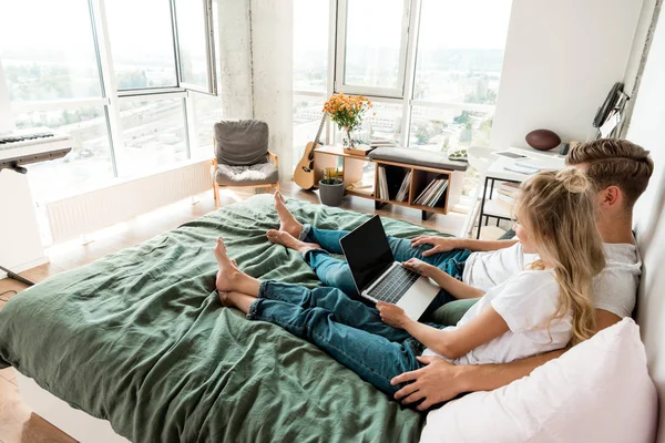 Vue latérale du jeune couple avec ordinateur portable numérique avec écran vierge reposant sur le lit à la maison — Photo de stock
