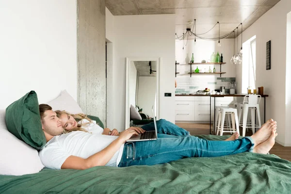 Vue latérale du jeune couple avec ordinateur portable numérique reposant sur le lit à la maison — Photo de stock