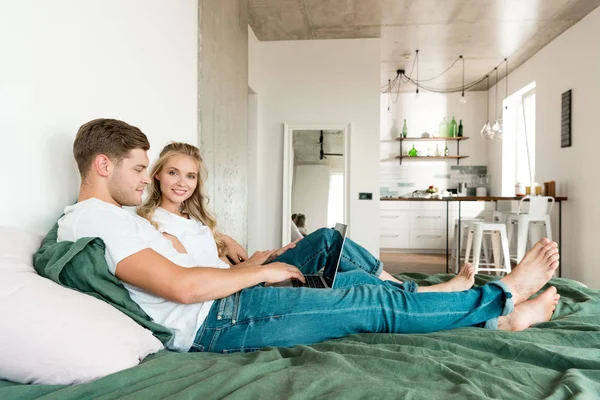 Sorrindo jovem casal com laptop digital descansando na cama em casa — Fotografia de Stock