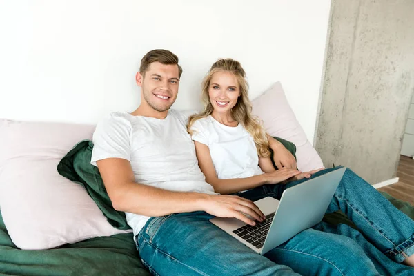 Улыбающаяся молодая пара с цифровым ноутбуком, лежащая на кровати дома — стоковое фото