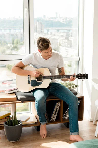 Молодой гитарист играет на акустической гитаре, сидя у окна дома — стоковое фото