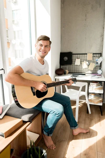 Молодой улыбающийся мужчина играет на акустической гитаре дома — стоковое фото