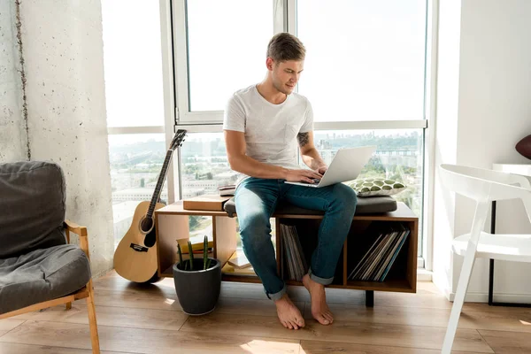 Молодой человек в повседневной одежде с ноутбуком, сидя у окна дома — стоковое фото
