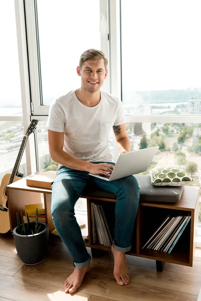 Hombre sonriente en ropa casual con portátil sentado en la ventana en casa - foto de stock