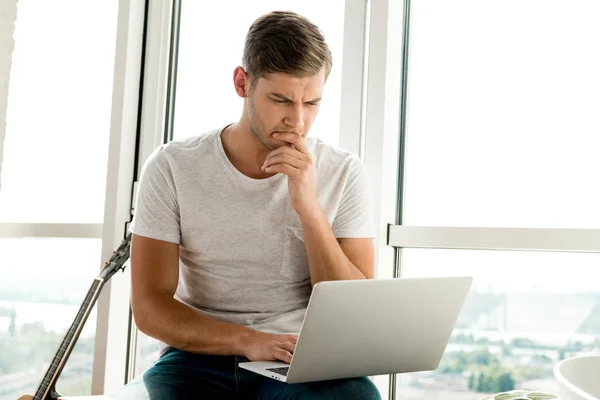 Homem focado em roupas casuais usando laptop enquanto sentado na janela em casa — Fotografia de Stock