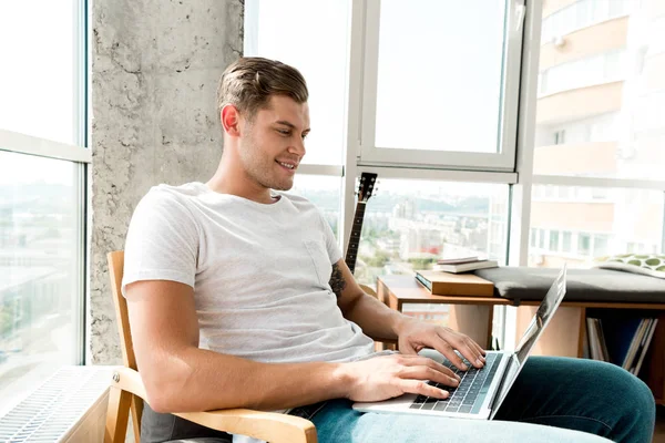 Hombre sonriente en sillón usando el ordenador portátil en casa - foto de stock