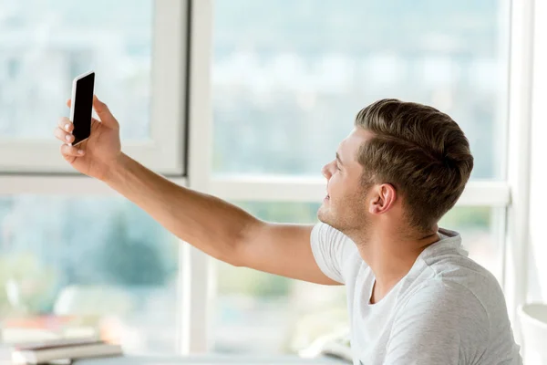 Вид збоку молодого чоловіка, який бере селфі на смартфон вдома — стокове фото