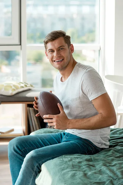 Hombre alegre con pelota de rugby en las manos mirando a la cámara en casa - foto de stock