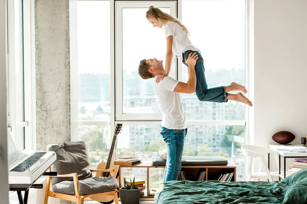 Side view of cheerful man holding girlfriend in hands while standing at window at home — Stock Photo