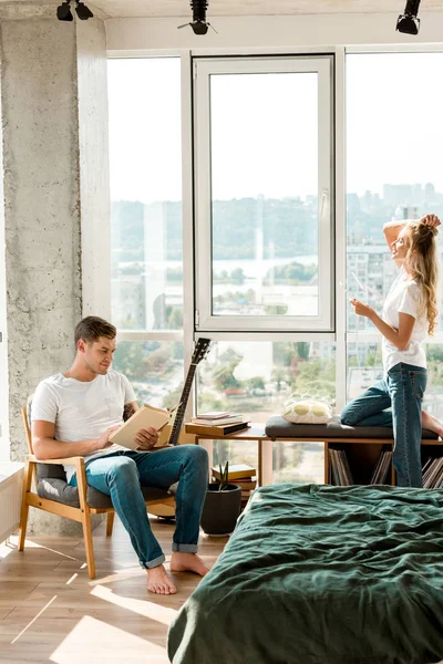 Young woman in earphones listening music while boyfriend reading book in armchair at home — Stock Photo