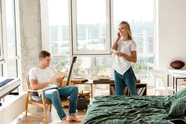 Young woman in earphones listening music while boyfriend reading book in armchair at home — Stock Photo