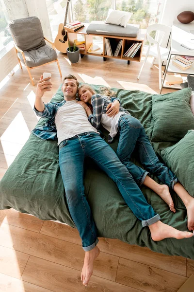 Vista de ángulo alto de alegre pareja joven tomando selfie en el teléfono inteligente mientras está acostado en la cama en casa - foto de stock