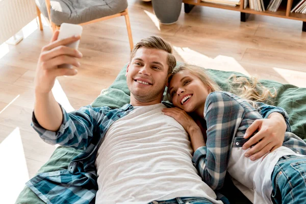 Retrato de alegre pareja joven tomando selfie en el teléfono inteligente mientras está acostado en la cama en casa - foto de stock