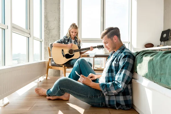 Hombre enfocado usando tableta digital mientras su novia toca la guitarra acústica en casa - foto de stock