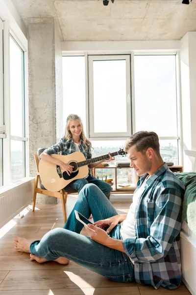 Junger Mann nutzt digitales Tablet, während Freundin zu Hause Akustikgitarre spielt — Stockfoto