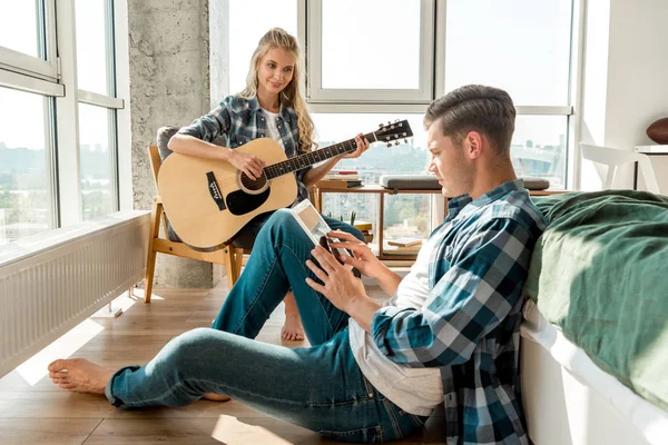 Mann nutzt digitales Tablet, während Freundin zu Hause Akustikgitarre spielt — Stockfoto