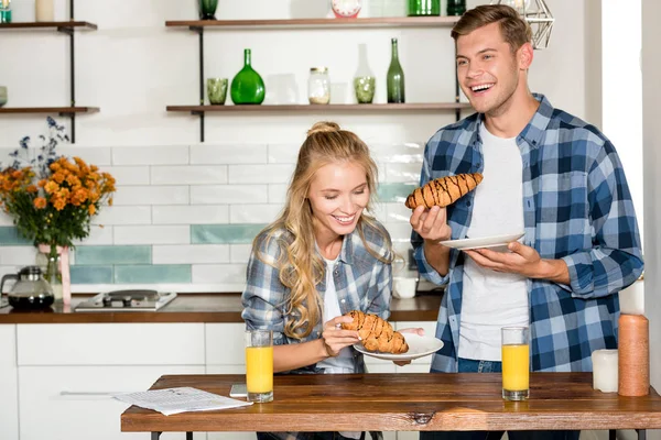 Portrait de couple heureux mangeant des croissants pour le petit déjeuner dans la cuisine à la maison — Photo de stock
