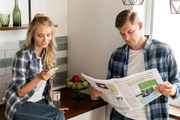 Casal com smartphone e jornal passando a manhã na cozinha em casa — Fotografia de Stock