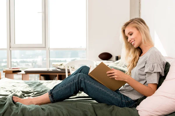 Vista lateral de sorrir bela mulher ler livro na cama em casa — Fotografia de Stock