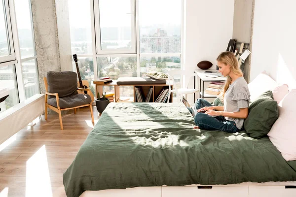 Vue latérale de belle femme en utilisant un ordinateur portable tout en étant assis sur le lit à la maison — Photo de stock