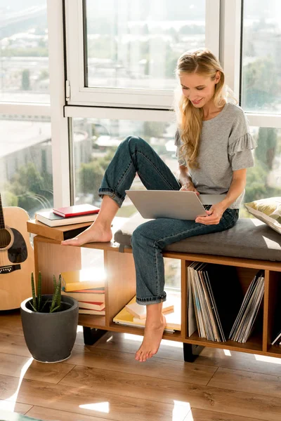 Улыбающаяся молодая женщина с помощью ноутбука, сидя у окна дома — стоковое фото