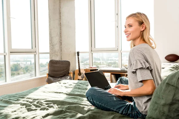 Belle femme souriante avec ordinateur portable assis sur le lit à la maison — Photo de stock