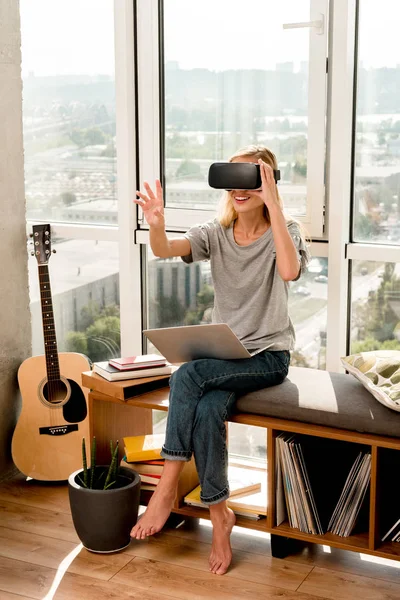 Mujer joven en auriculares vr con portátil sentado en la ventana en casa - foto de stock